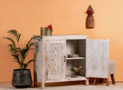 Side angle image with open door of a white washed cabinet handcrafted from mango wood with engraved doors; artisanal Indian mango wood furniture, 90 × 90 × 40 cm.